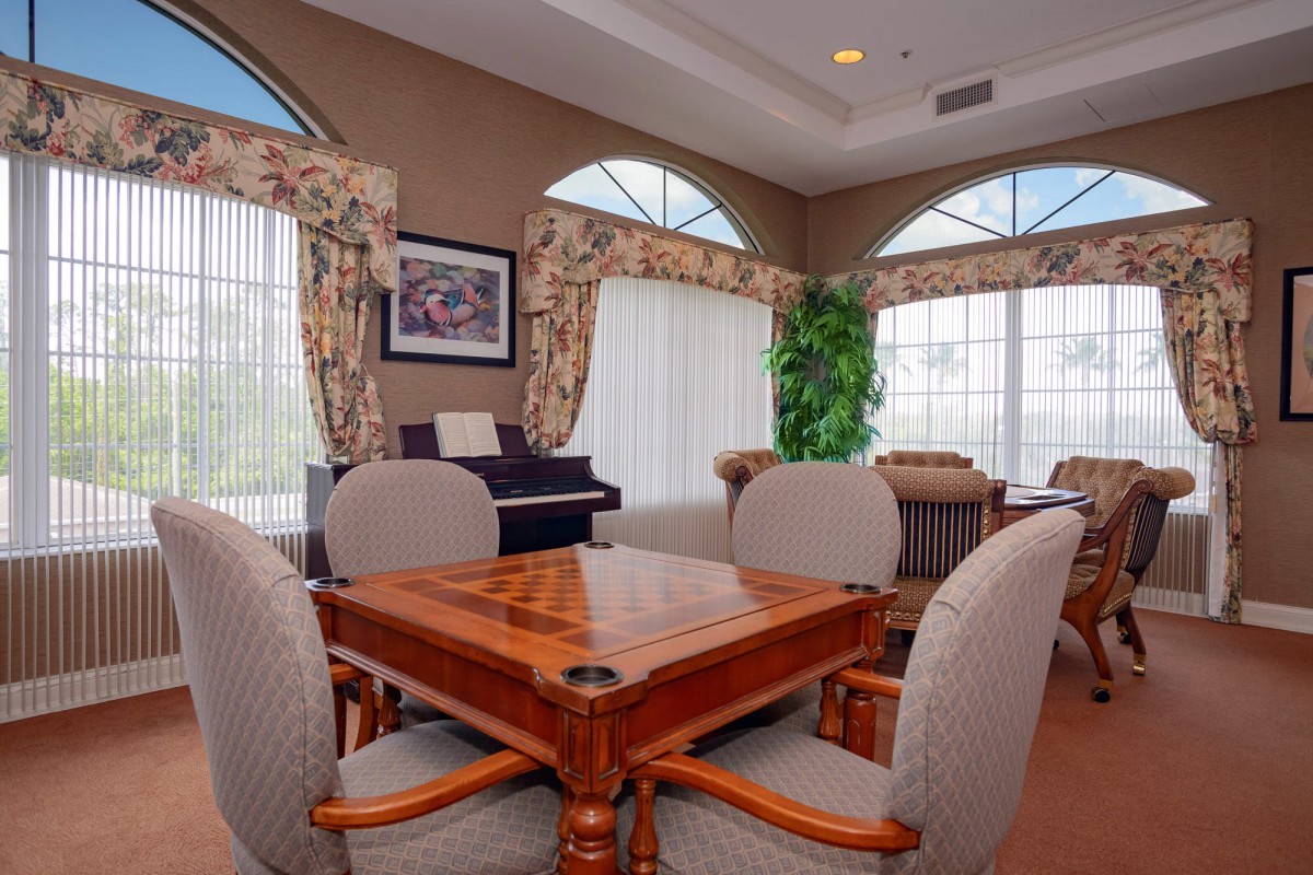 A cozy living room with a central wooden table surrounded by chairs, featuring decorative curtains and a piano.