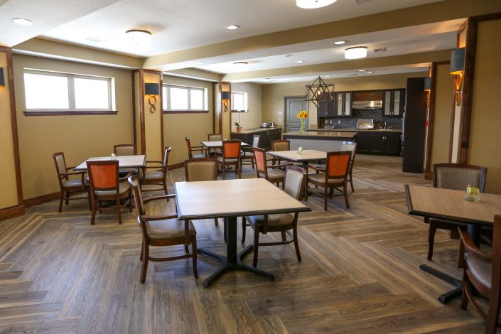 A spacious dining area with multiple tables and chairs, featuring a kitchen area in the background and wood flooring.