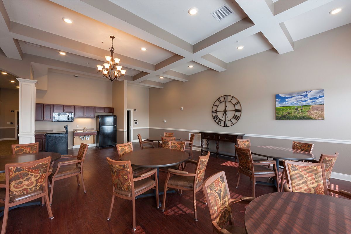 A spacious dining room featuring round tables and woven chairs, with a large wall clock and chandelier adding elegance to the decor.