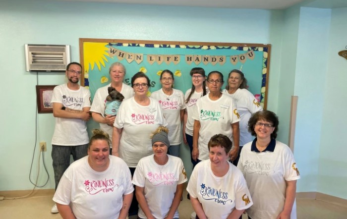 A group of people posing together indoors, wearing matching t-shirts in front of a bulletin board.