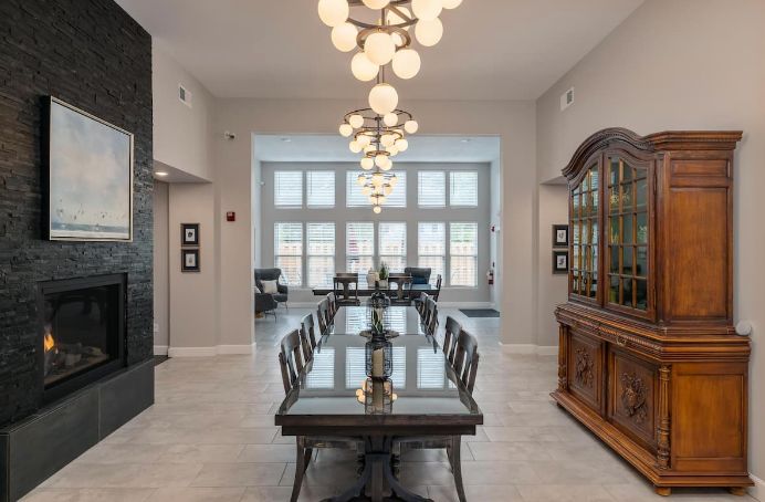 A spacious dining room featuring a long table with chairs, illuminated by modern chandeliers and natural light streaming in through large windows.