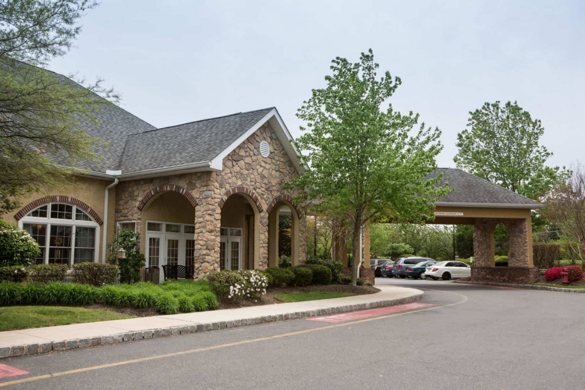 A modern building with a stone facade and an entrance surrounded by trees and a parking area.