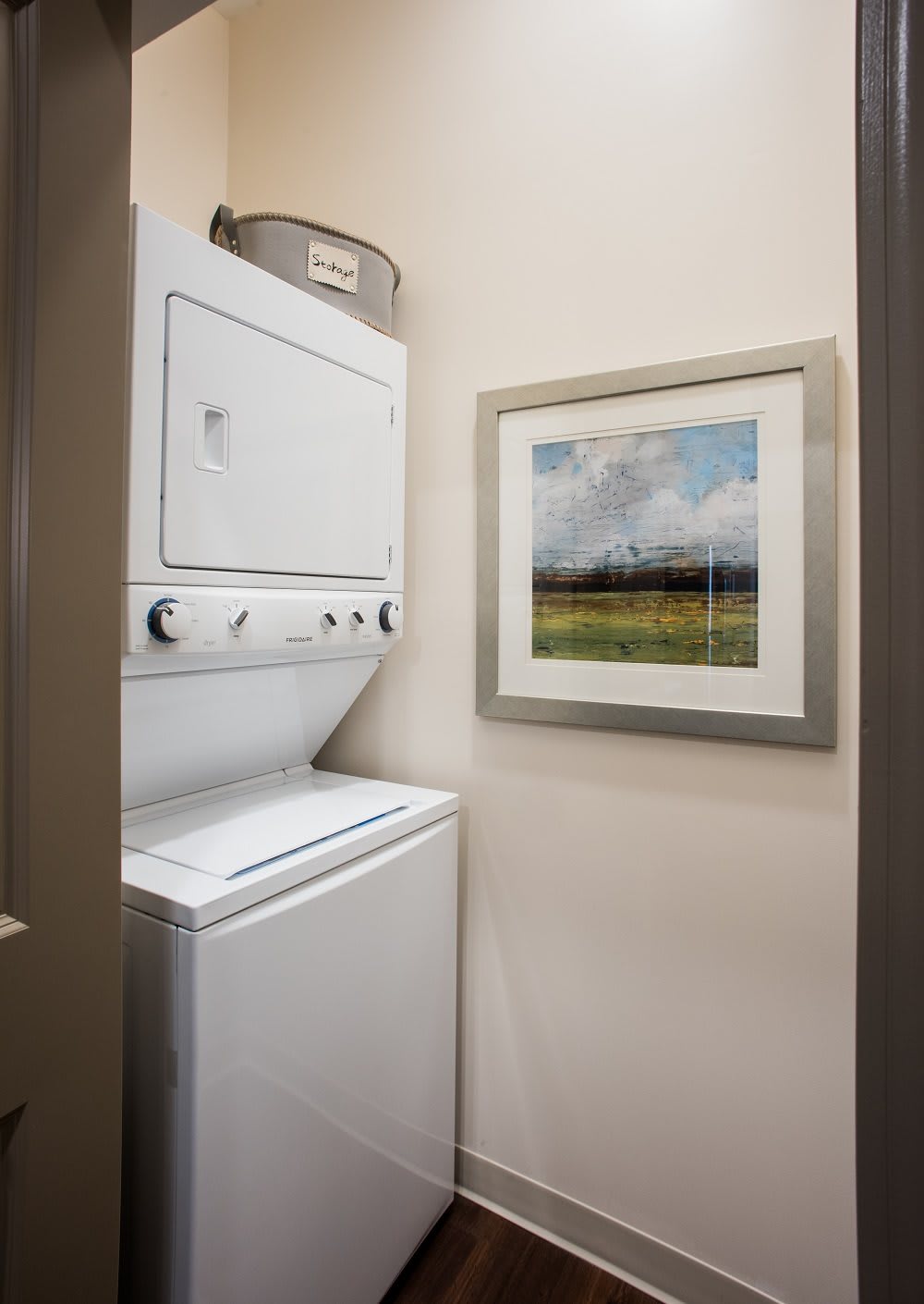 A small laundry area featuring a stacked washer and dryer unit with a framed landscape painting on the wall.