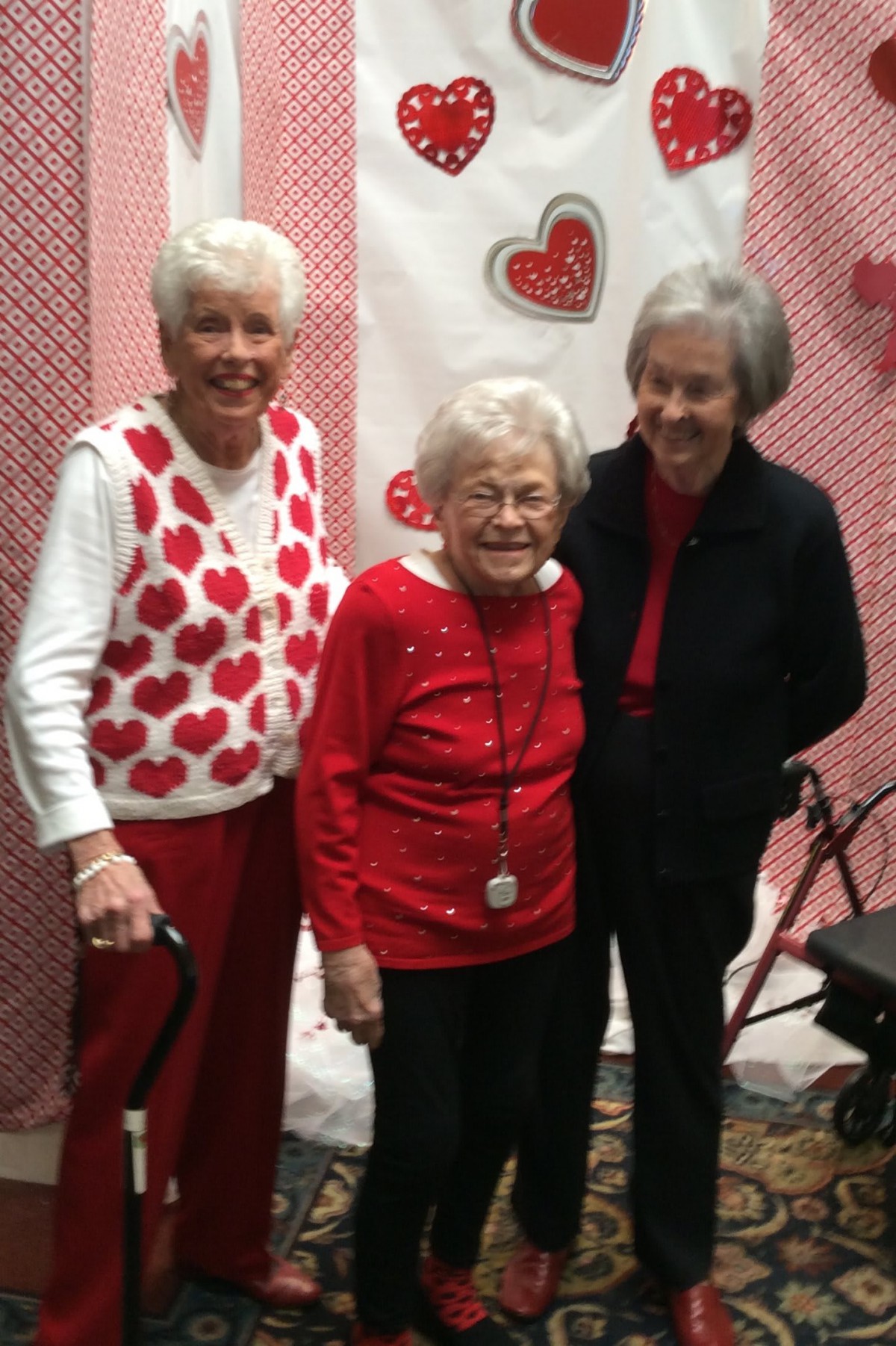 Three elderly women pose together in front of heart-themed decorations, all dressed in red and festive attire.