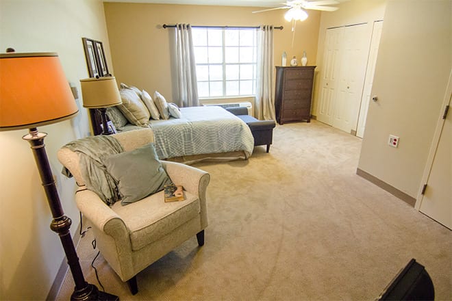 A cozy bedroom with a bed, comfortable chair, and natural lighting from a large window.
