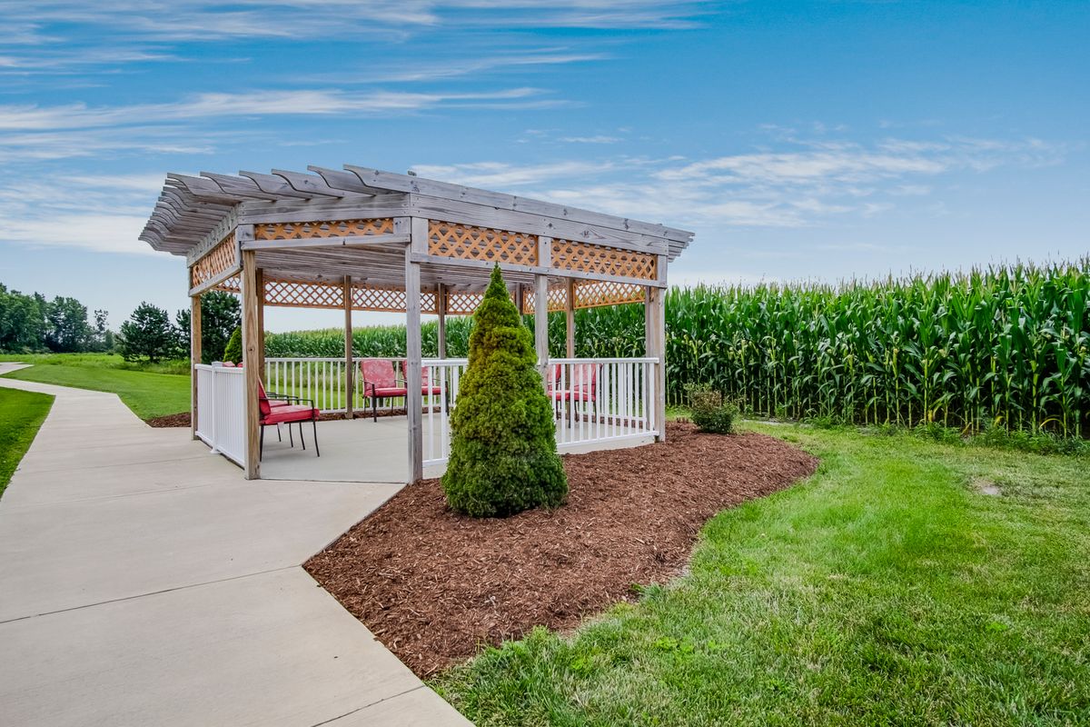 A charming gazebo in a rural setting surrounded by greenery and a cornfield under a blue sky.