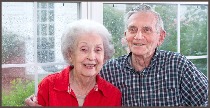 An elderly couple smiles warmly while sitting together by a window.