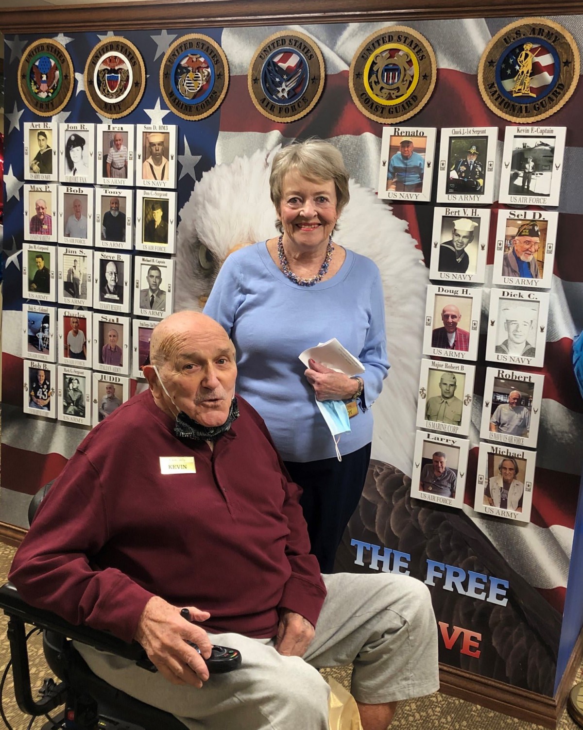 An elderly man in a wheelchair and a woman standing beside him, with a background displaying military portraits and symbols.