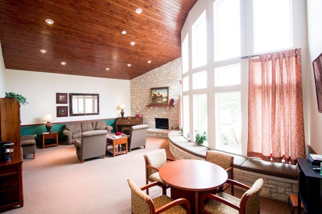 A well-lit living room featuring a cozy arrangement of sofas and chairs around a fireplace, with large windows and a wooden ceiling enhancing the inviting atmosphere.