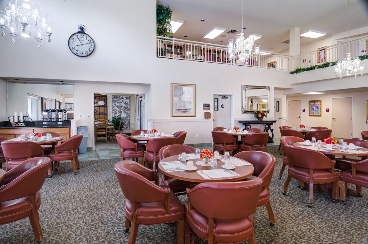 A well-lit indoor dining area with round tables and cushioned chairs, featuring a carpeted floor and a large wall clock.