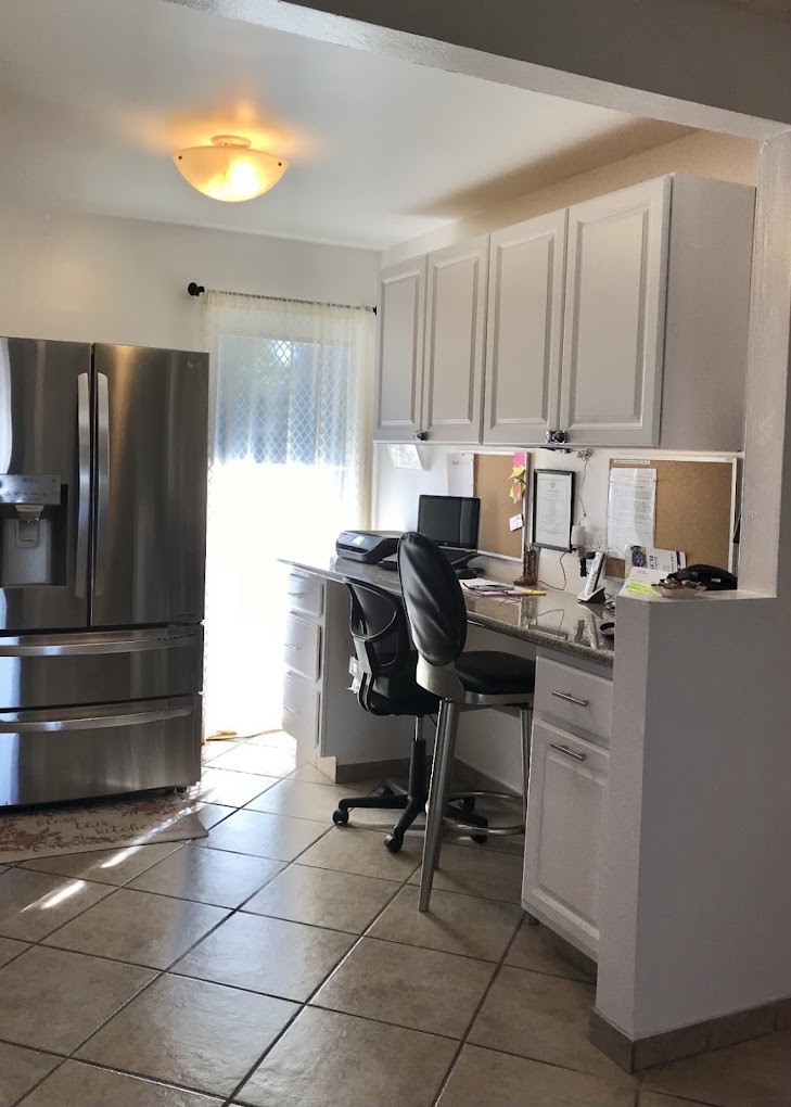 A modern kitchen with an integrated home office area, featuring white cabinets, a stainless steel refrigerator, and a desk with a computer.