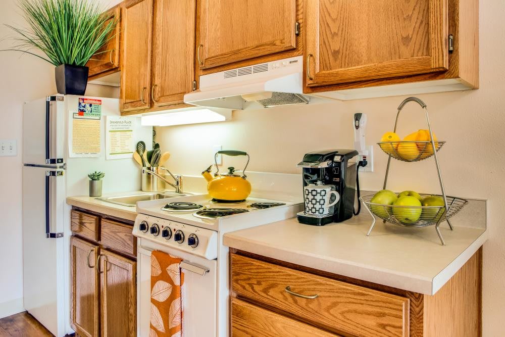 A cozy kitchen with wooden cabinets, featuring a stove with a yellow teapot, a coffee maker, and a fruit basket on the countertop.