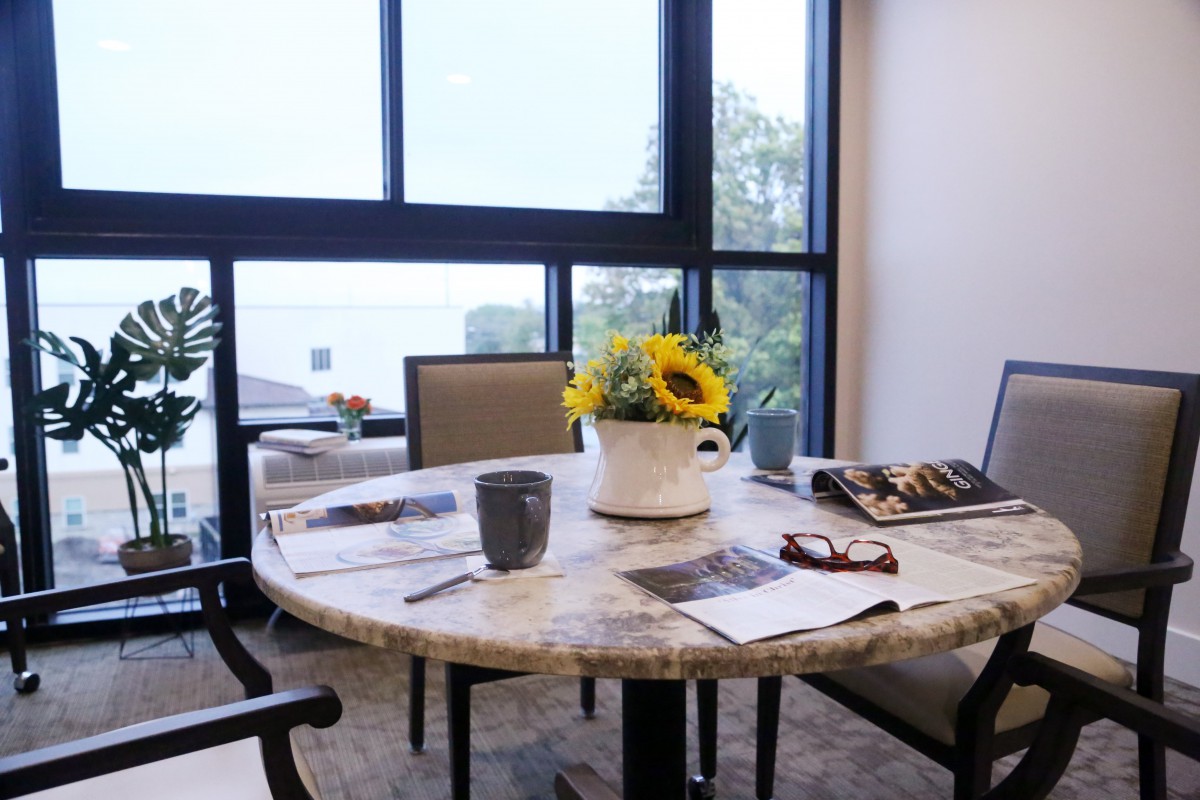 A cozy dining table by the window with a sunflower centerpiece, coffee mug, and newspapers.