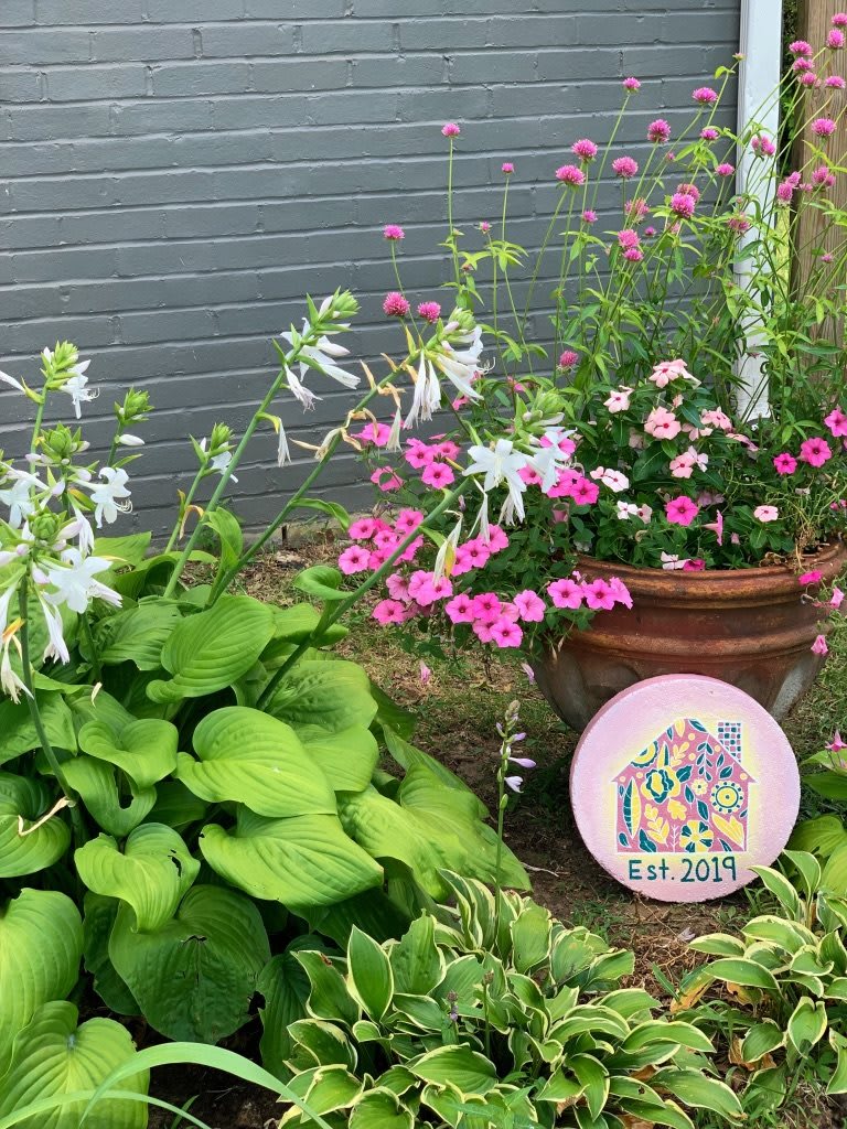 A vibrant garden scene featuring lush hostas and pink flowers in a terracotta planter against a brick wall.