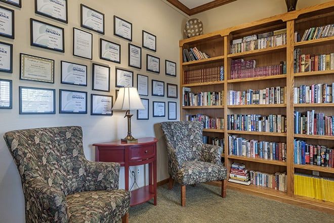 A cozy reading nook with bookshelves, a lamp, and two upholstered chairs, adorned with framed certificates on the wall.