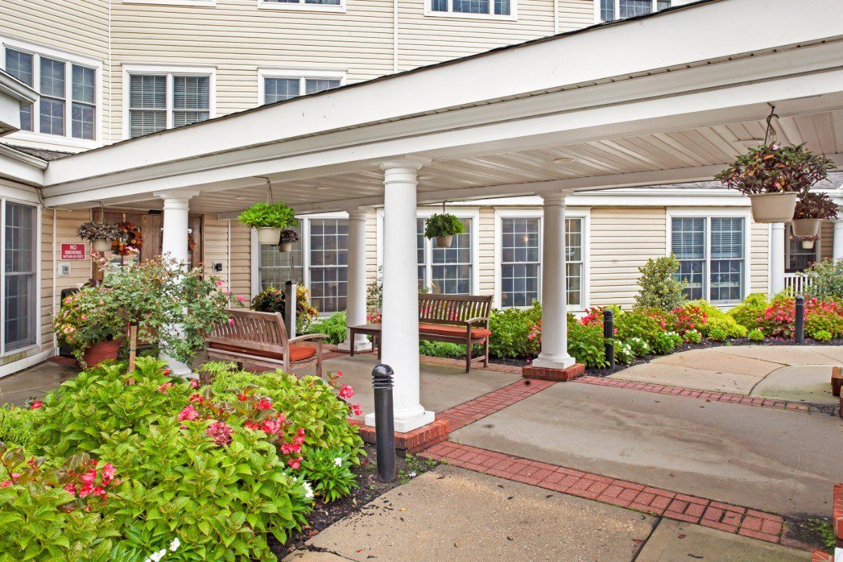 A covered entrance of a building with white columns, surrounded by flowers and benches.