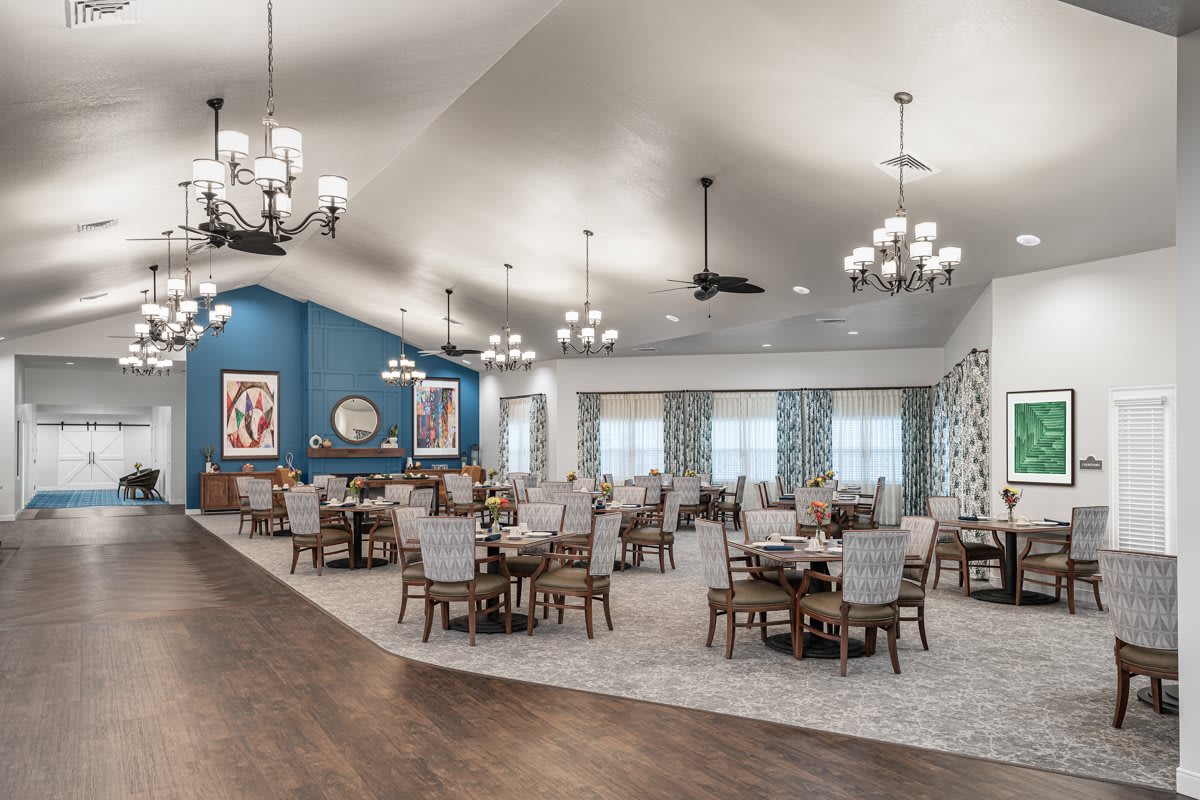 A spacious dining room with neatly arranged tables and chairs, illuminated by elegant chandeliers. A blue accent wall features decorative art, complementing the carpeted flooring.