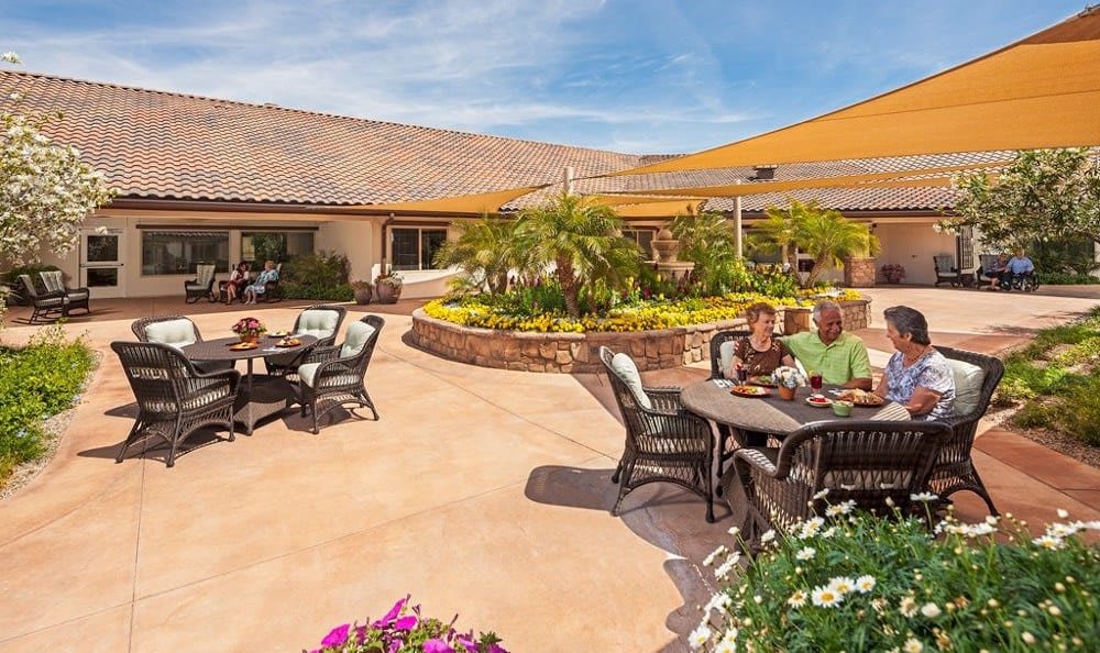 People enjoying a sunny day on a spacious outdoor patio with tables and chairs surrounded by vibrant flowers.