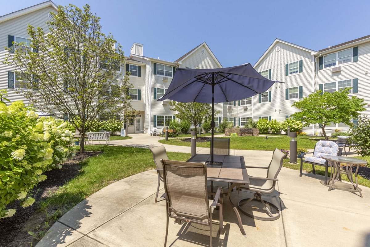 A serene outdoor patio area with a table and chairs under an umbrella, surrounded by a well-maintained garden in front of a residential apartment building.