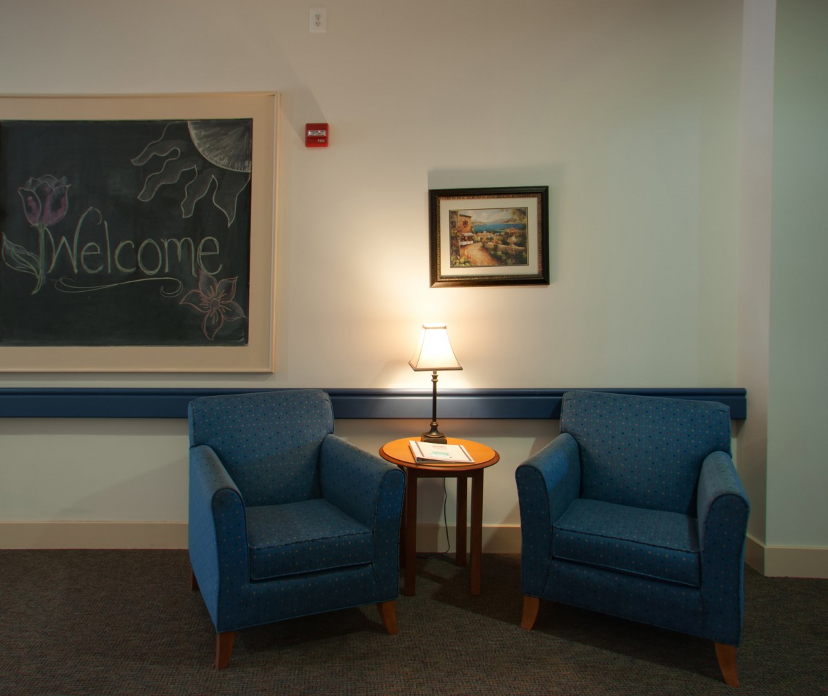 A cozy seating area with two blue armchairs, a small wooden table, and a lamp in between. A chalkboard and artwork decorate the wall.