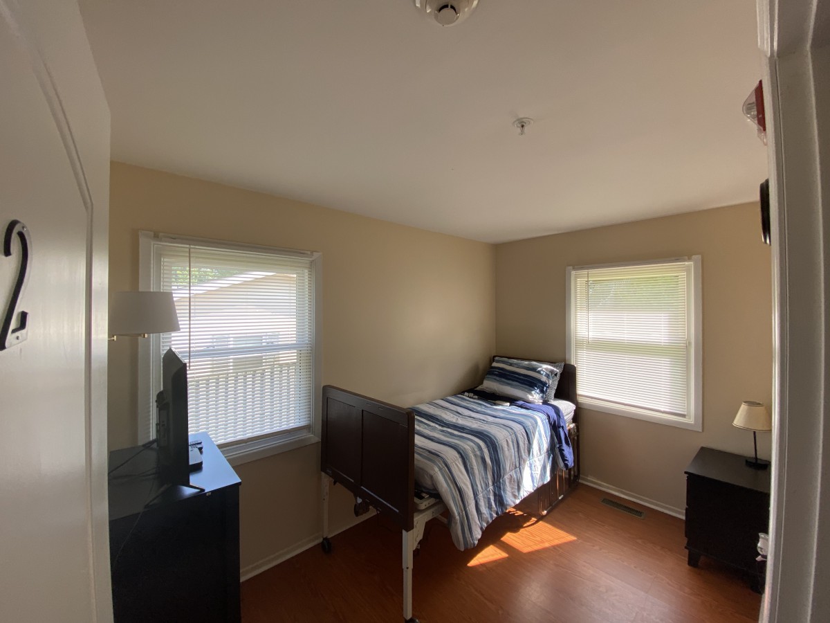 A simple bedroom with a single bed and two windows, furnished with a dresser and a lamp.