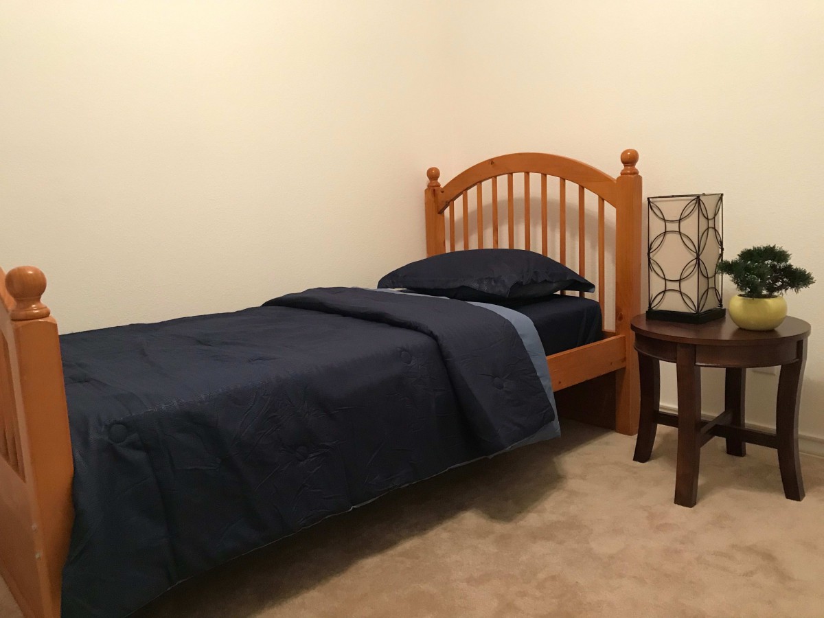 A simple minimalist bedroom with a single wooden bed, dark bedding, and a nightstand with a lamp.
