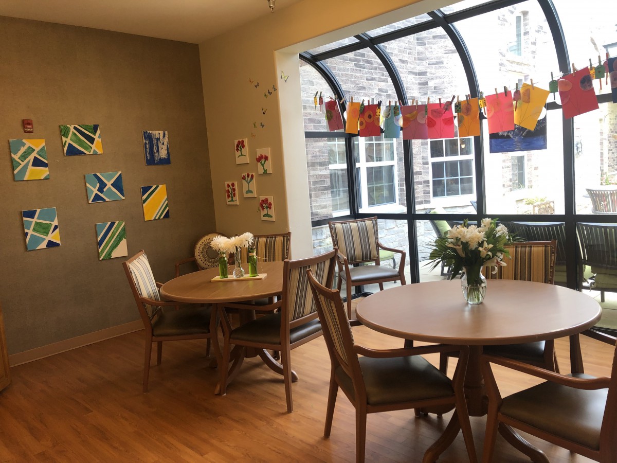 A cozy dining area adorned with colorful artwork and fresh flowers, featuring round tables and large windows.