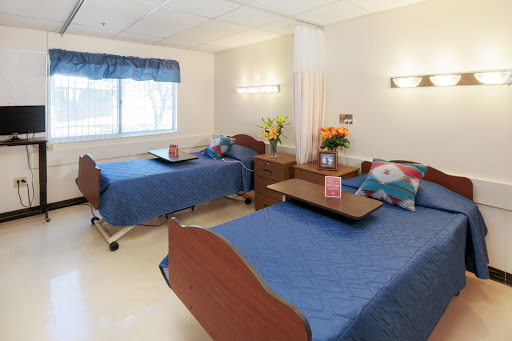 A well-lit shared patient room in a hospital with two beds, each equipped with blue bedding and bedside furniture.