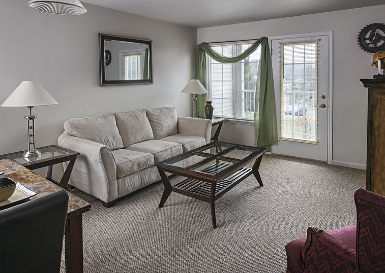 A cozy living room with a beige sofa, glass-top coffee table, and green curtains framing the window.
