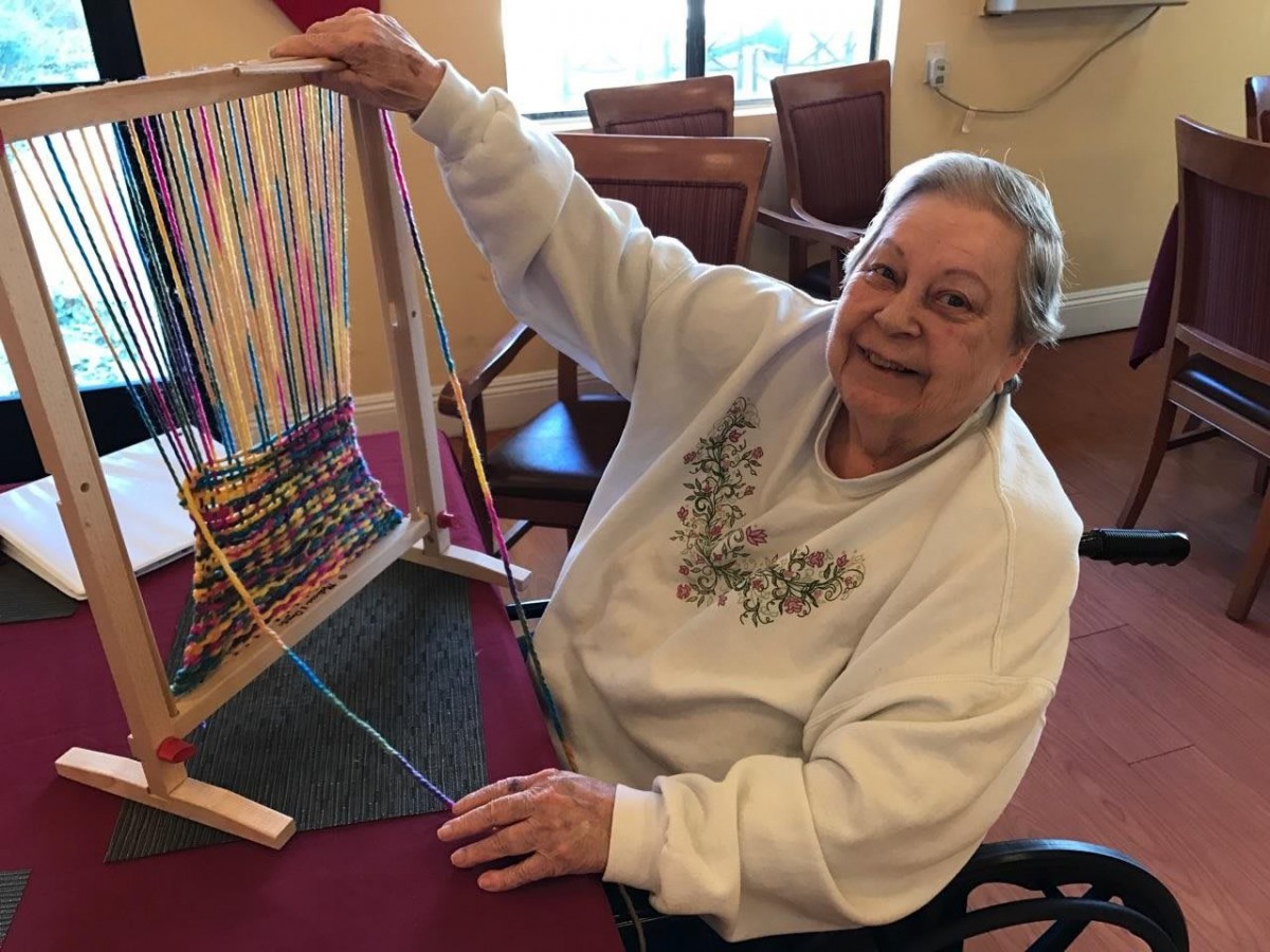 Elderly woman enjoys weaving on a loom, smiling while working on her craft project.