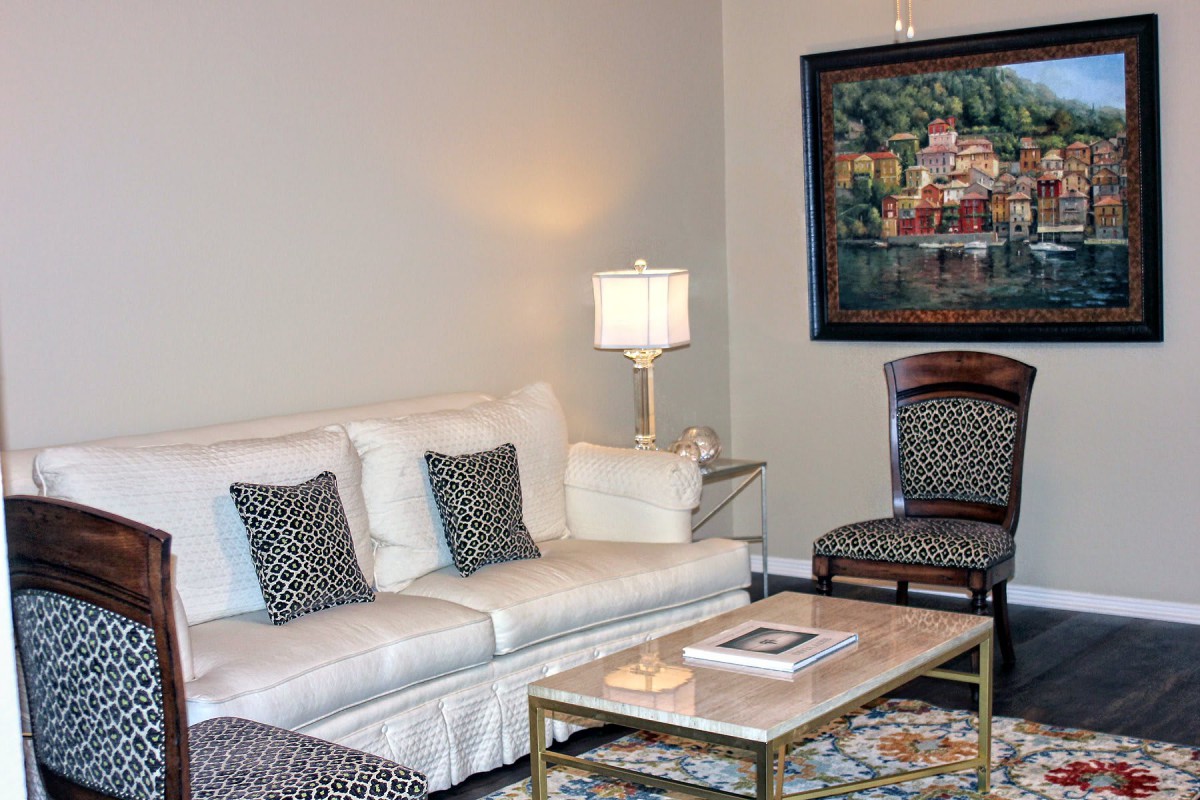 A cozy living room with a white sofa, patterned pillows, a coffee table, and a lamp next to framed artwork on the wall.
