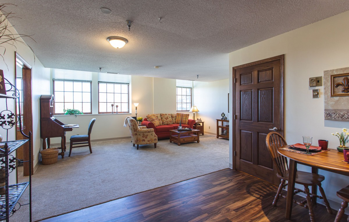 A spacious living room with a mix of carpet and wooden flooring, featuring comfortable seating and large windows.