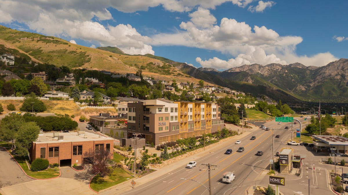 A scenic view of a mountainous urban area with a main road flanked by modern buildings under a partly cloudy sky.