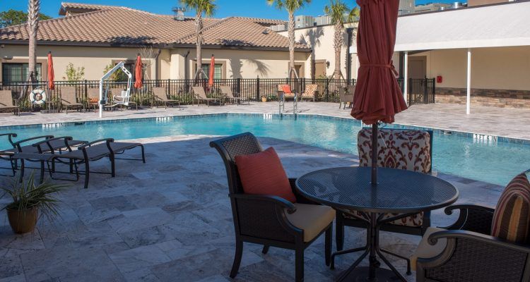 A sunny outdoor area featuring a swimming pool surrounded by patio furniture and umbrellas, set against a backdrop of palm trees in a residential complex.