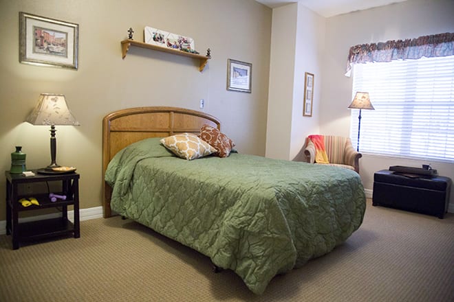 A cozy bedroom featuring a bed with a green quilt, wall art, and a standing lamp.