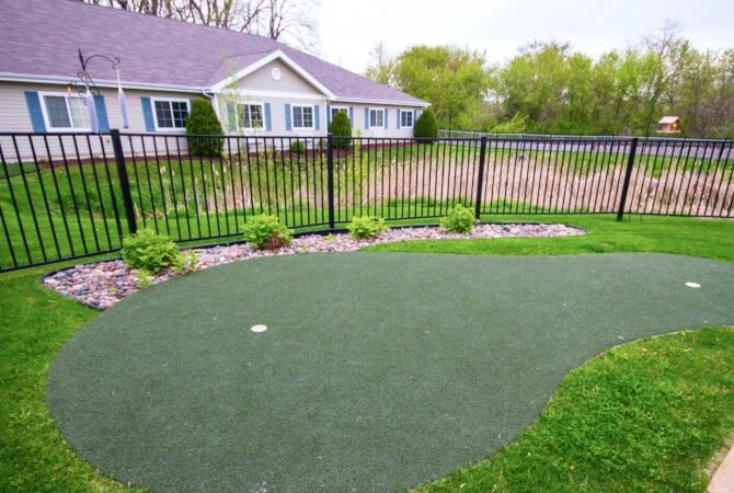 A backyard putting green with neatly landscaped surroundings and a view of a house.