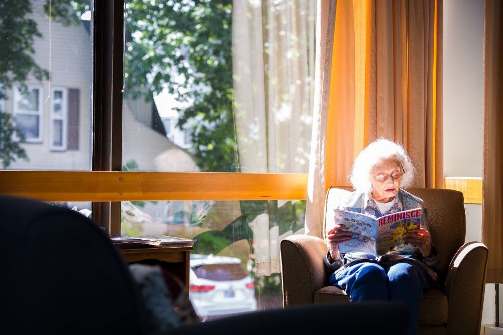 An elderly person reads a newspaper in a sunlit room by the window.