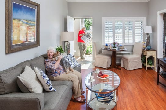 An elderly woman sits on a sofa with her pet dog, as a visitor enters the bright and cozy living room.