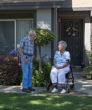 An elderly couple spending time together outside their house, with one person sitting in a wheelchair.