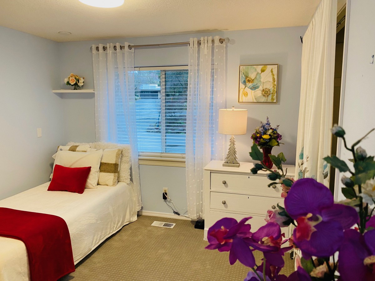 A cozy and bright bedroom with a white bed, accented by red pillows and blanket, featuring a dresser with a lamp and decorative flowers.