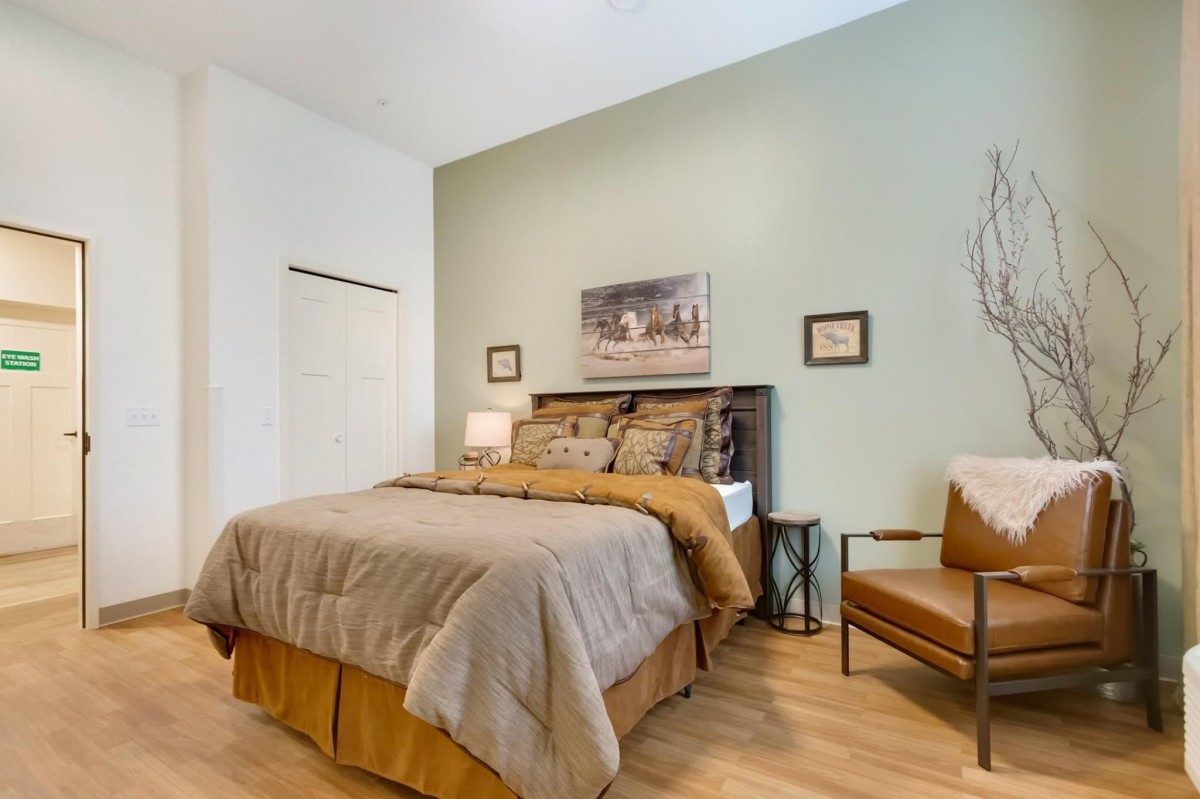 A cozy bedroom featuring a bed with neutral bedding, accompanied by a chair and decorative wall art.