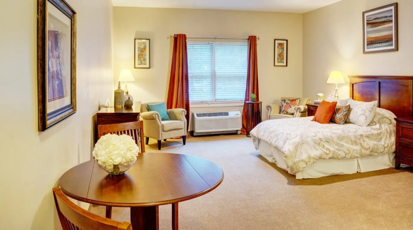 A cozy bedroom with warm lighting, featuring a bed, armchair, and a small table near a window with curtains.