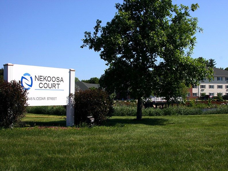 A sign for Nekoda Court stands in a grassy area with a tree and building in the background.