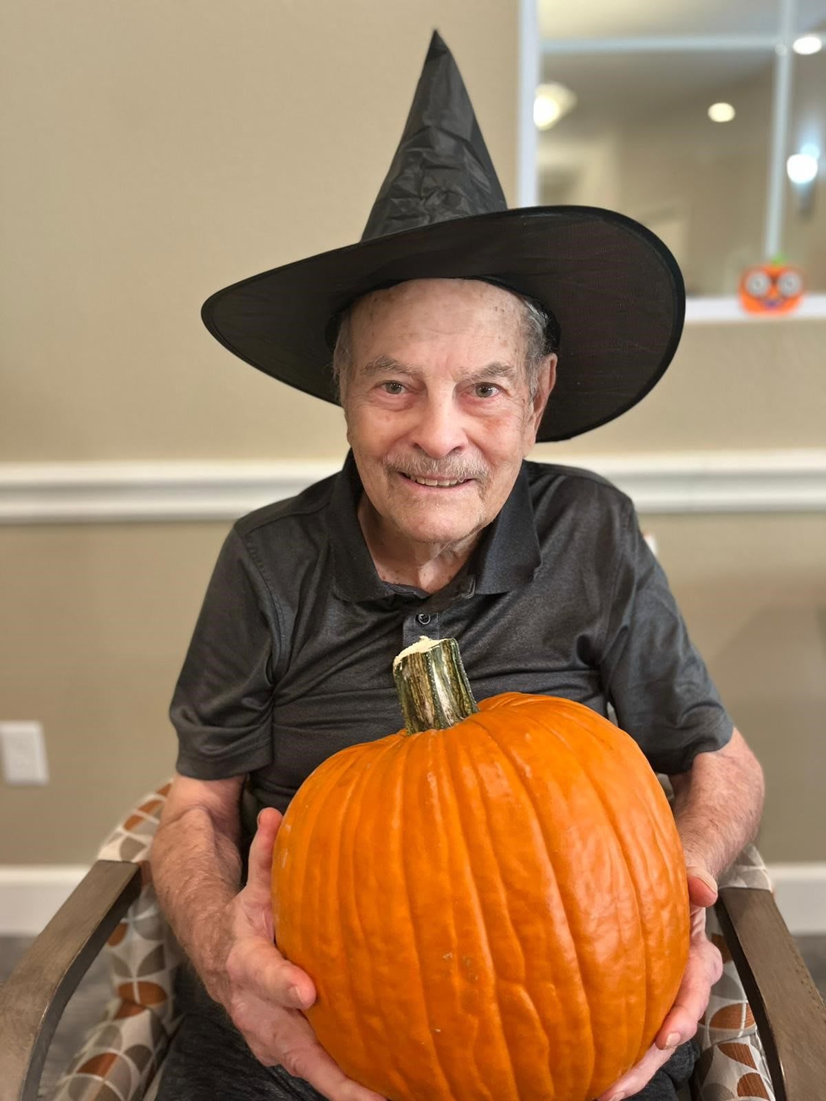 A man wearing a witch hat smiles while holding a large pumpkin.