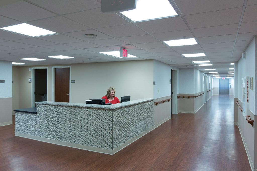 A hospital reception area with a staff member at the desk and a long corridor extending in the background.