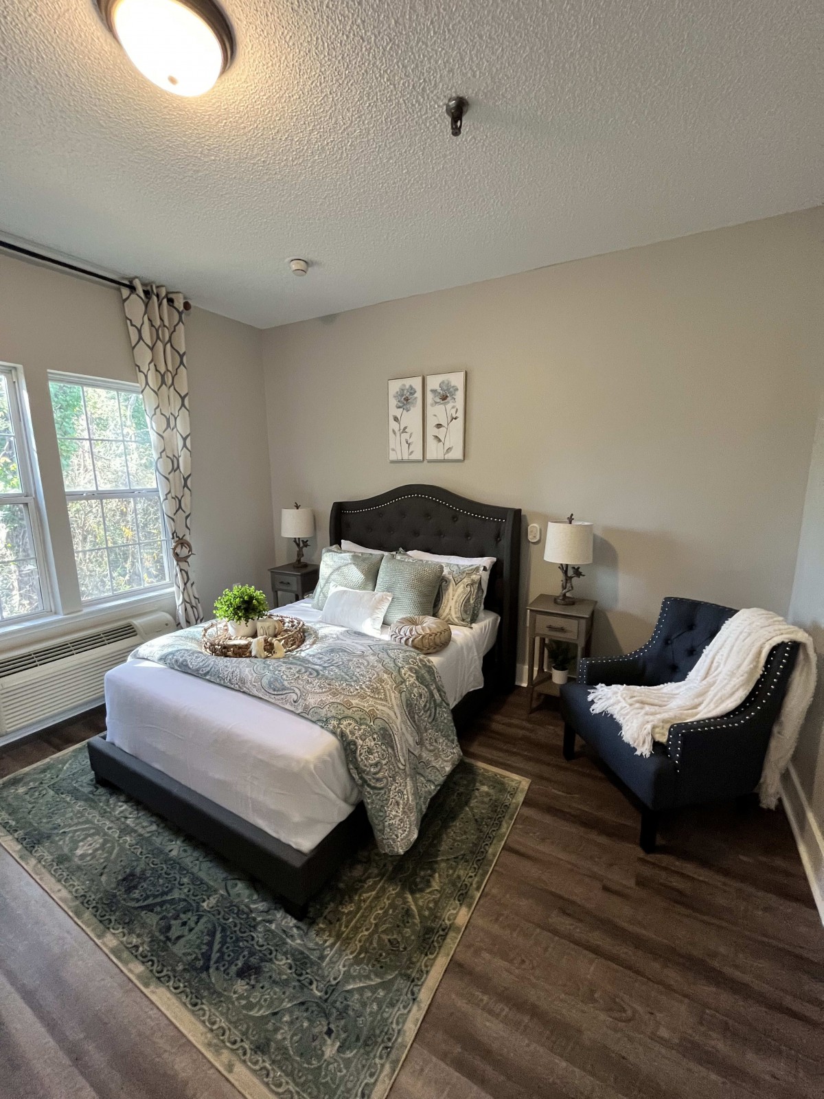 A cozy and well-decorated bedroom featuring a bed with pillows and blankets, an armchair, bedside lamps, a window with curtains, and a rug on the floor.