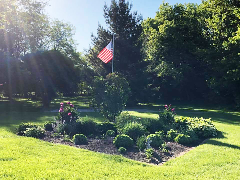 A sunlit garden with an American flag surrounded by lush greenery.