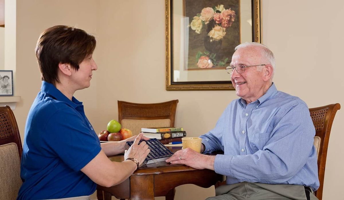 A caregiver converses with an elderly man at a table in a cozy room.