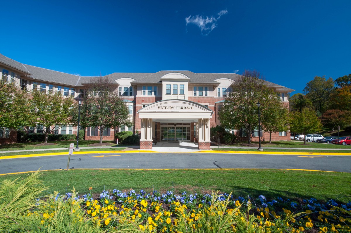 A stately brick building with a welcoming entrance, framed by vibrant flowers and clear blue skies.