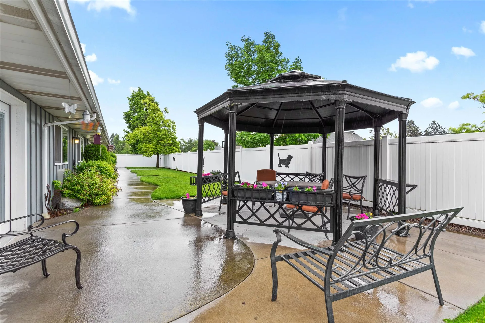 A backyard featuring a metal gazebo with seating, surrounded by a lush green lawn and a paved pathway.